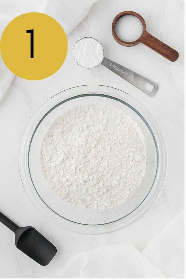 Flour in a glass mixing bowl with measuring spoons of baking powder and salt alongside.