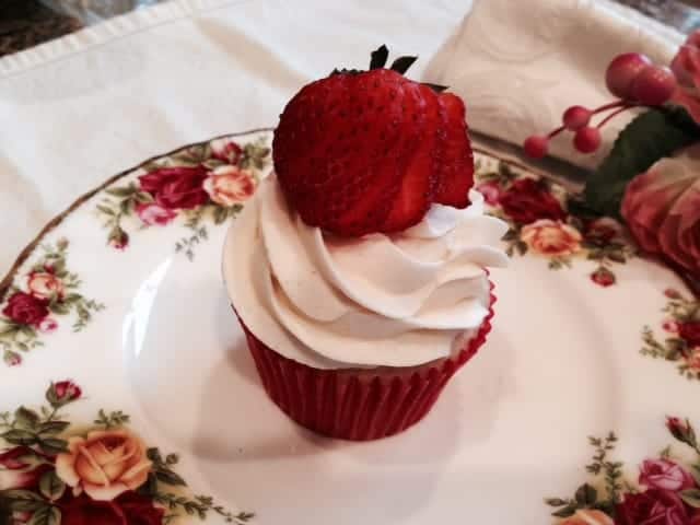 A strawberry mousse filled cupcake with a tall swirl of vanilla frosting and a fresh strawberry, on a rose patterned plate.