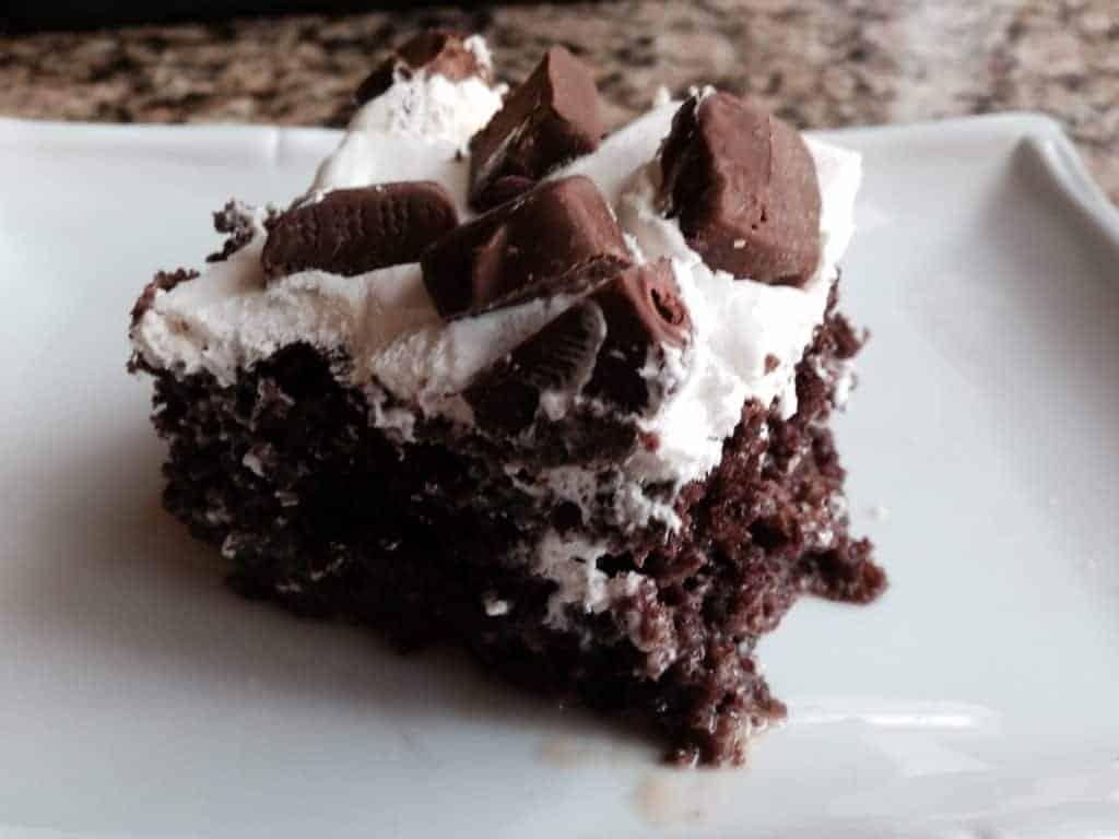 A slice of Milky Way poke cake on a white plate, showing the moist chocolate crumb under whipped topping and chopped candy bars.
