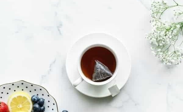A white teacup of tea on a saucer with lemon slices and blueberries on a marble table.