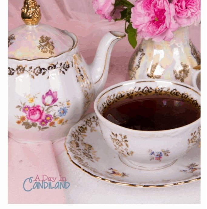 A gold trimmed floral teapot and a cup of tea beside a vase of pink roses.