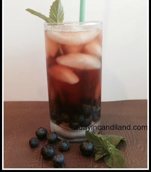 A tall glass of blueberry mint iced tea over ice with a sprig of mint and a straw.