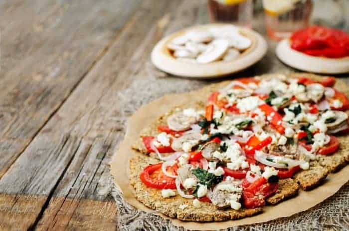 Chia seed pizza on parchment topped with tomatoes, mushrooms, red peppers and crumbled cheese.