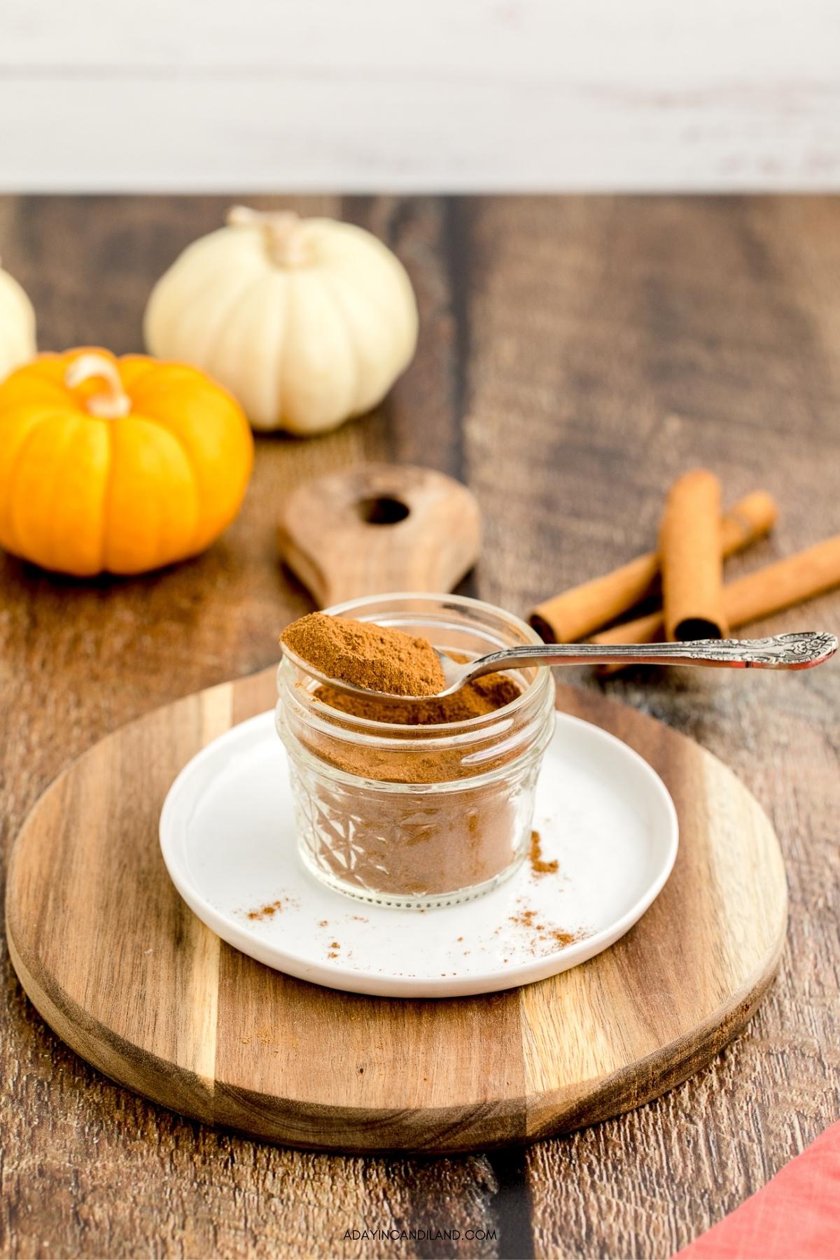 Jar of pumpkin pie spice with a spoon on top and cinnamon sticks on the table. 