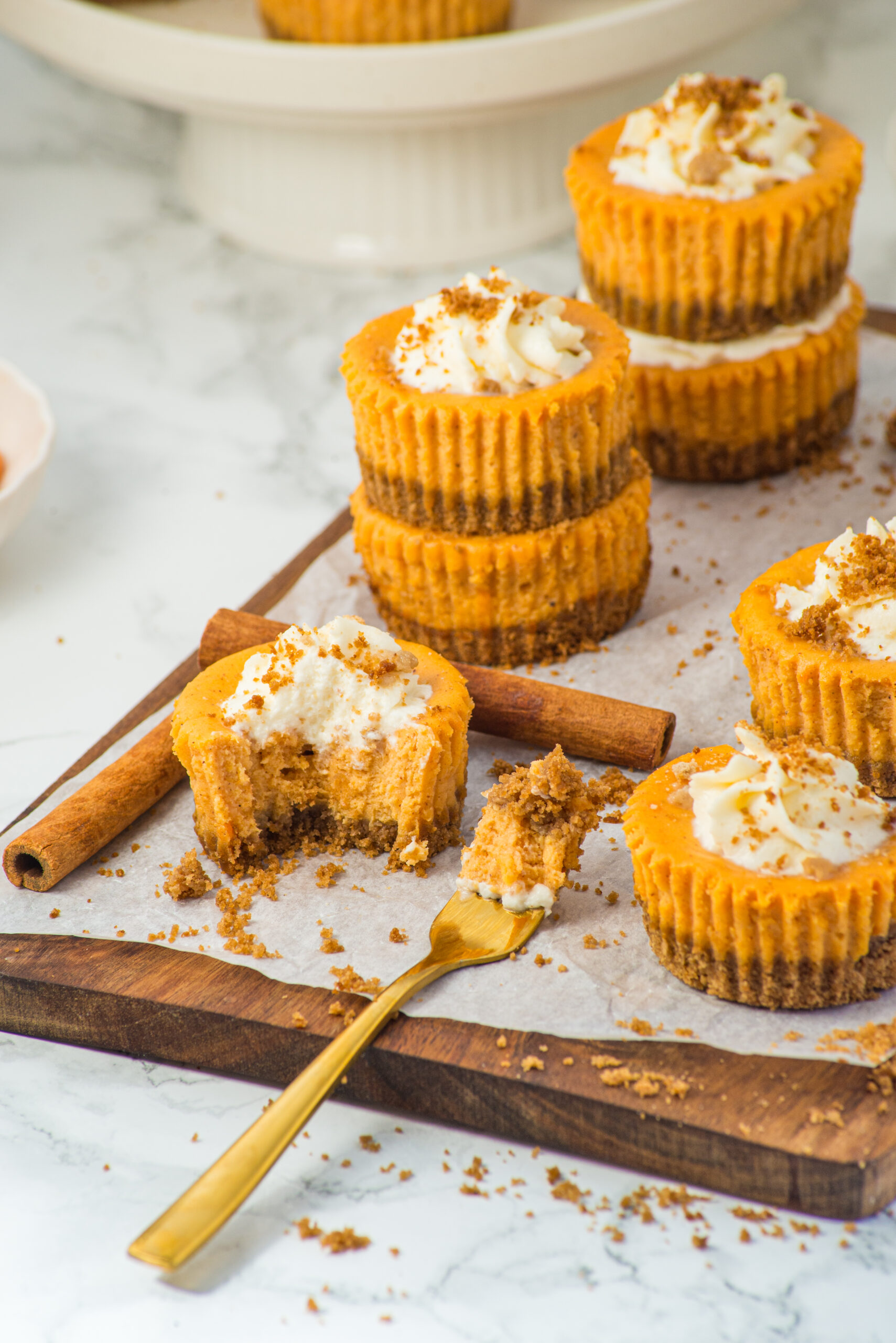 Stacked mini Pumpkin Cheesecakes on a wood cutting board with cinnamon sticks and fork with bite on it. 