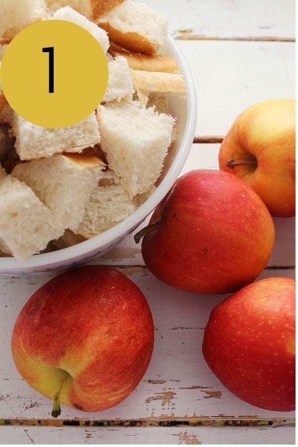 Cubed French bread in a white bowl with fresh red apples on a wooden table.