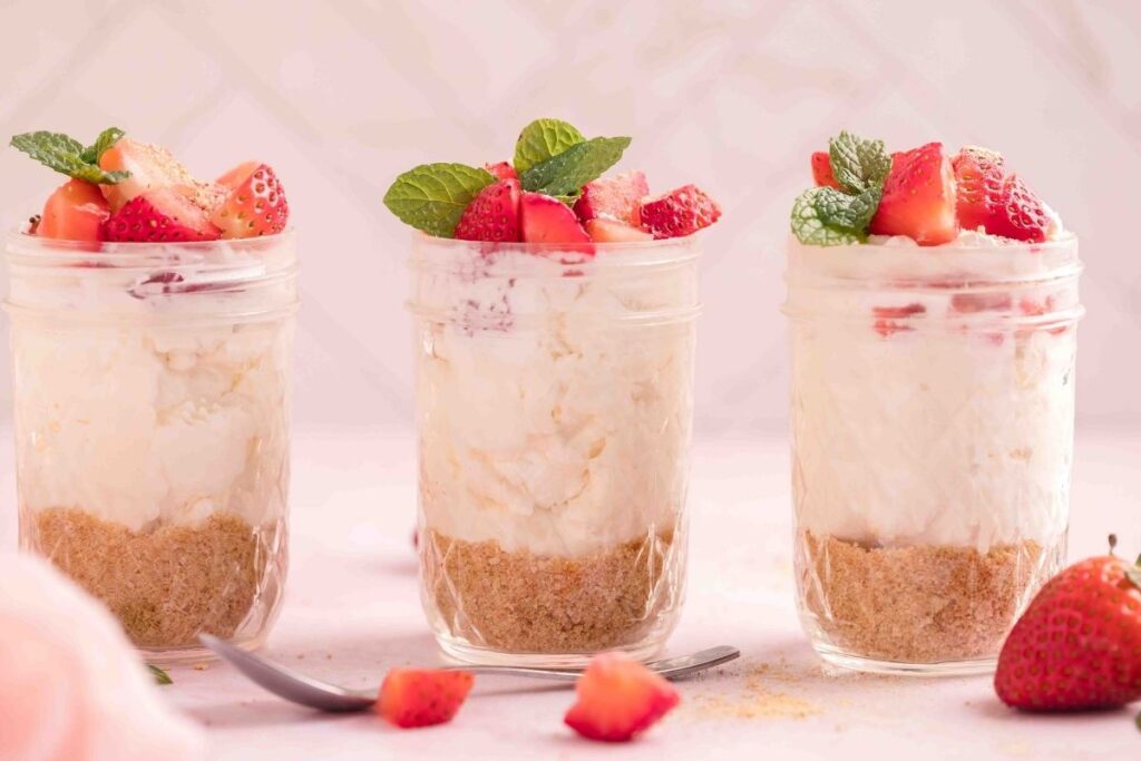 3 individual jars of Strawberry Cheesecakes with some sliced strawberries on the table.