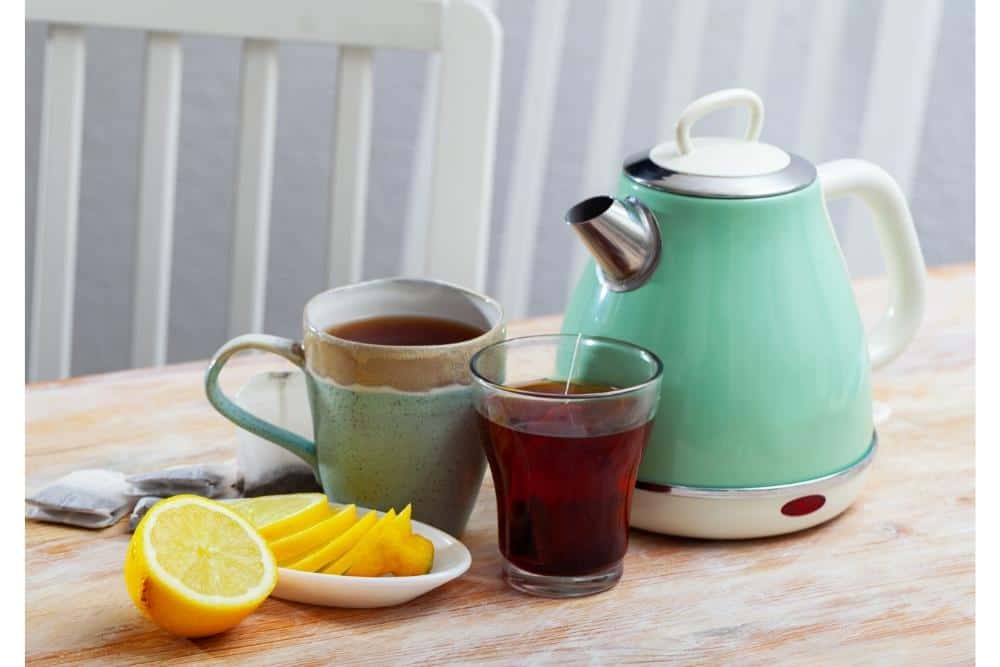 A mint green electric tea kettle on its base beside cups of tea and lemon slices.