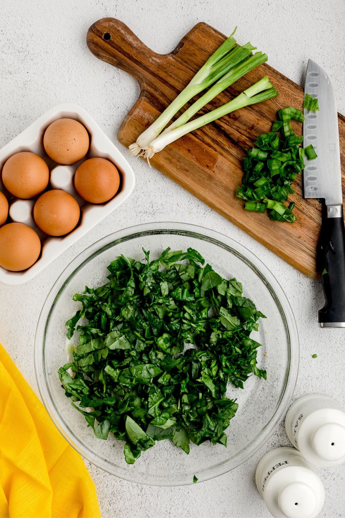 Chopped fresh spinach in a glass bowl with green onions and eggs on a wooden board.