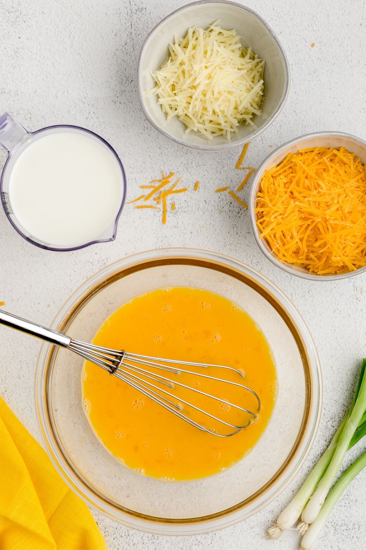 Six eggs whisked smooth in a glass bowl beside bowls of cheddar and Parmesan.
