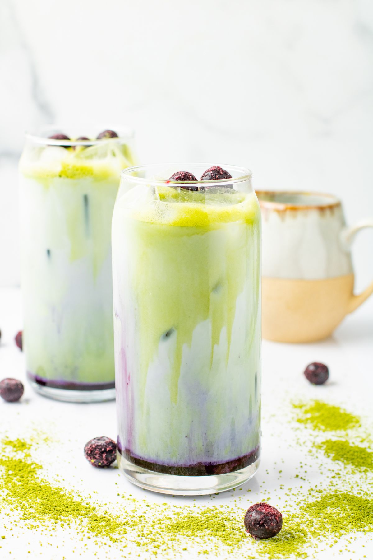 Two glasses of Blueberry Matcha Latte. with a coffee cup in the back. 