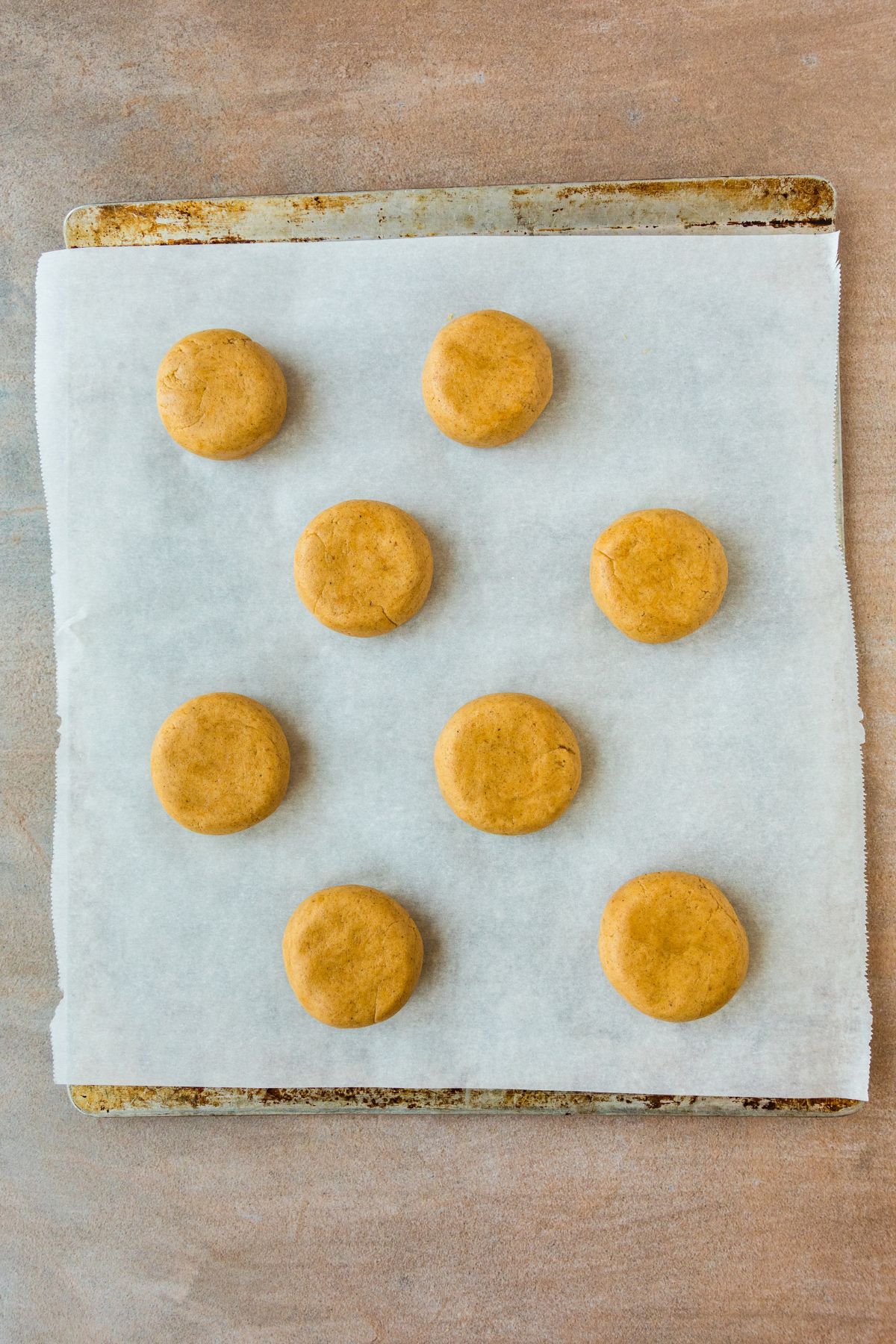 Eight balls of pumpkin cookie dough pressed flat on a parchment lined baking sheet.