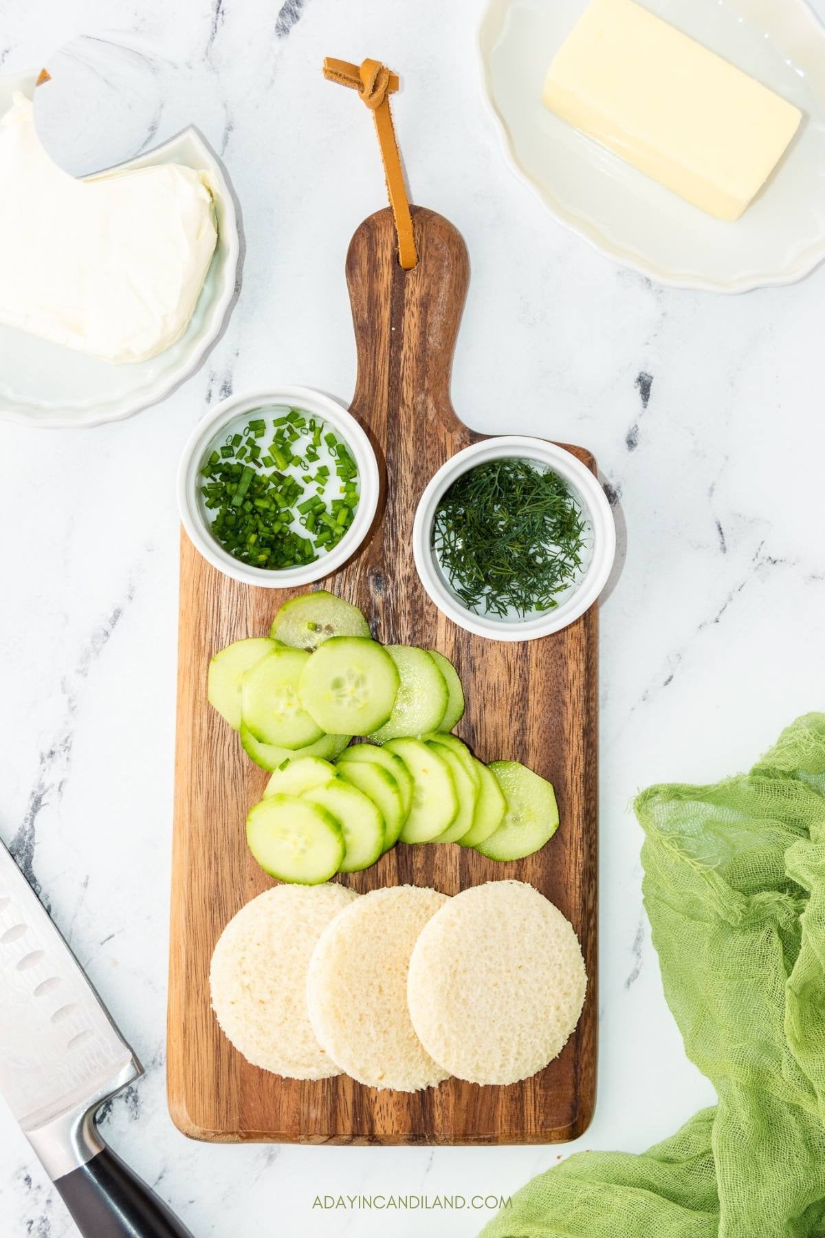Sliced cucumbers, bread rounds, dill and chives on a wooden board.