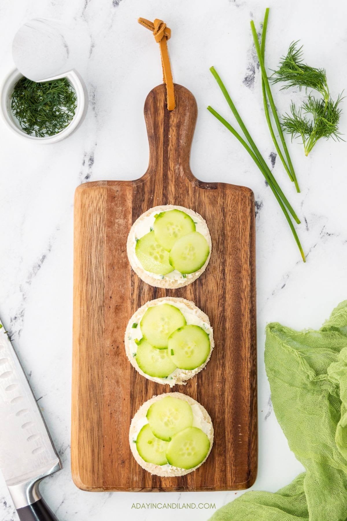 Finished cucumber tea sandwiches topped with dill on a wooden board.