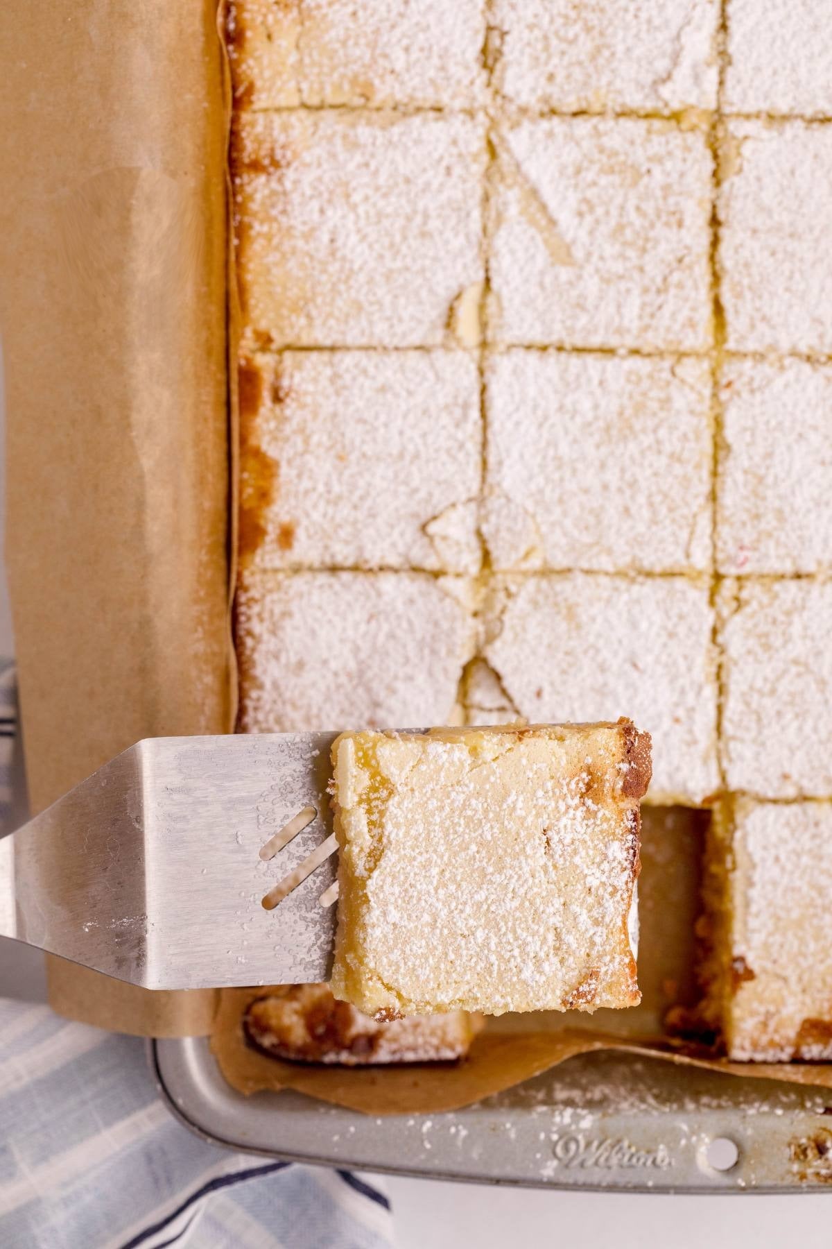 Baked lemon bars dusted with powdered sugar, cut into squares, one lifted on a spatula.