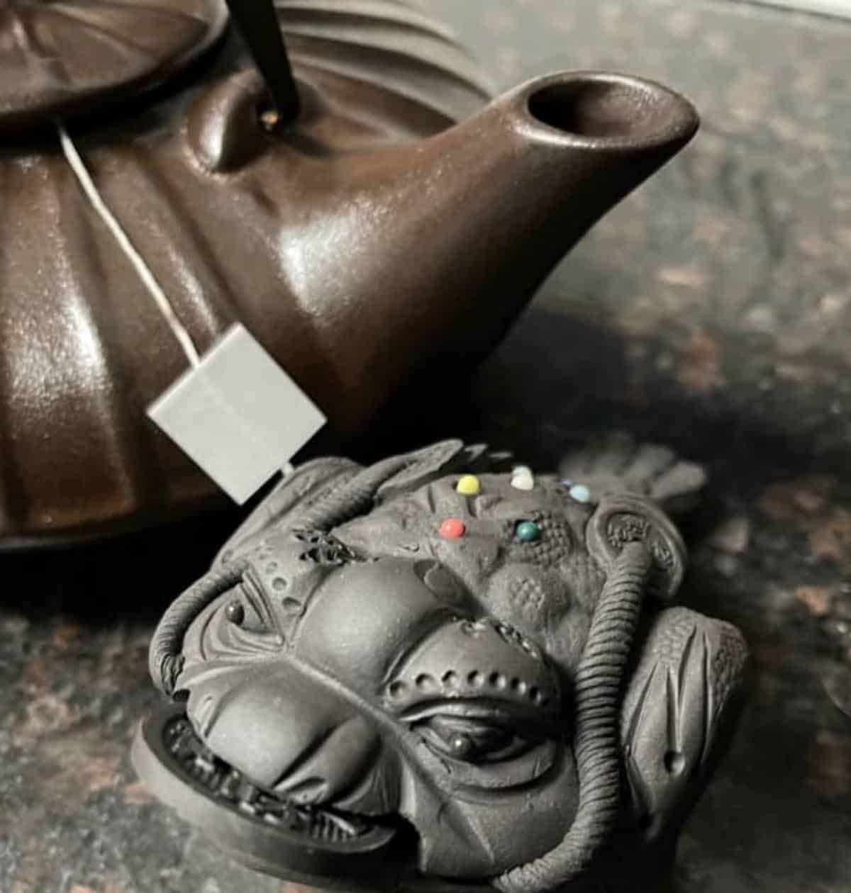 A dark clay toad tea pet sitting beside a brown clay teapot.