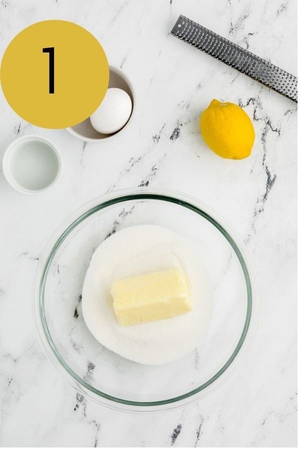 Softened butter and sugar in a glass mixing bowl with an egg, lemon and zester nearby.