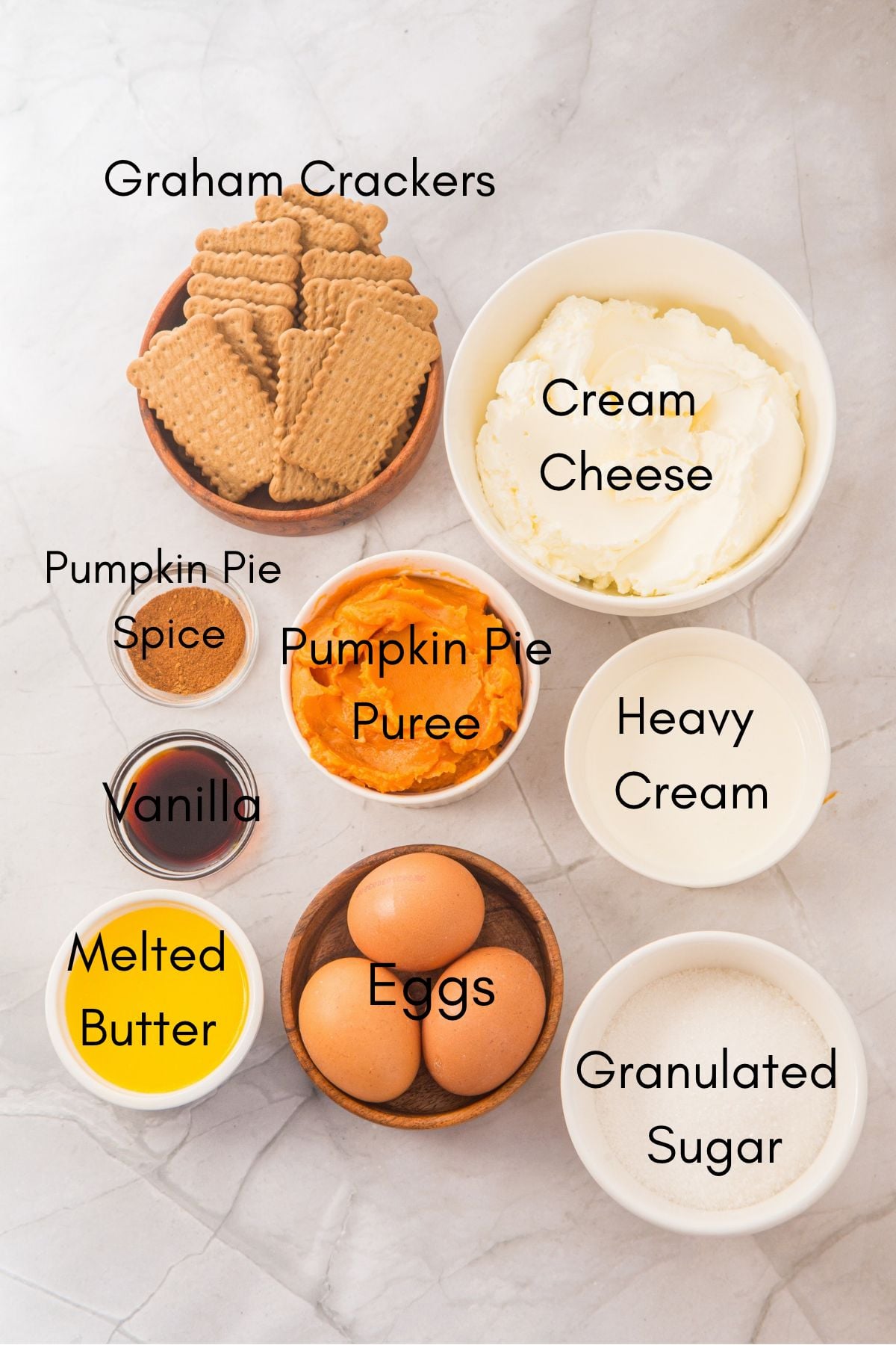 Labeled ingredients for mini pumpkin cheesecakes: graham crackers, cream cheese, pumpkin pie spice, pumpkin puree, heavy cream, vanilla, melted butter, eggs and granulated sugar.