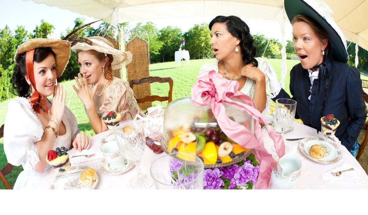 Friends in wide brimmed straw hats and a dark brimmed hat laughing around a garden tea party table.