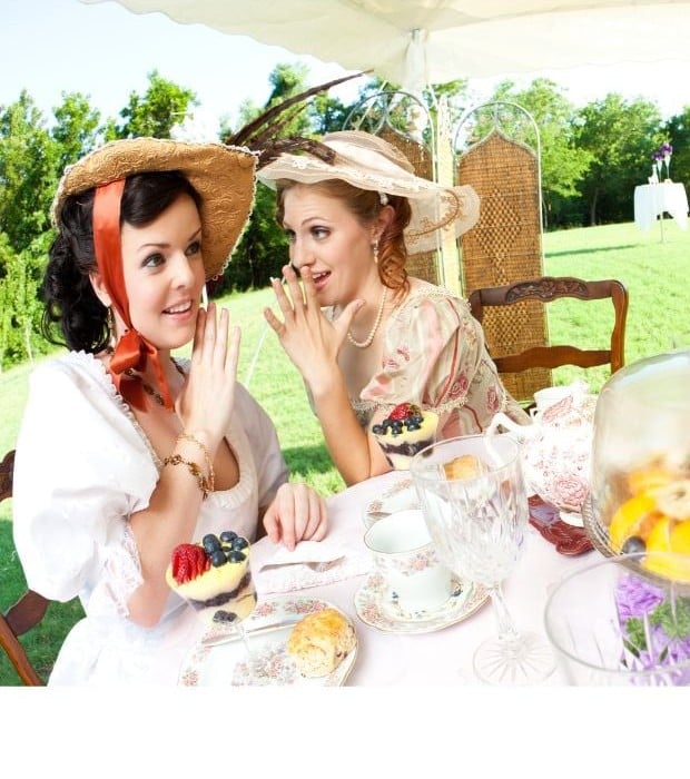 Two women in wide brimmed straw hats with ribbons and flowers enjoying tea and dessert.
