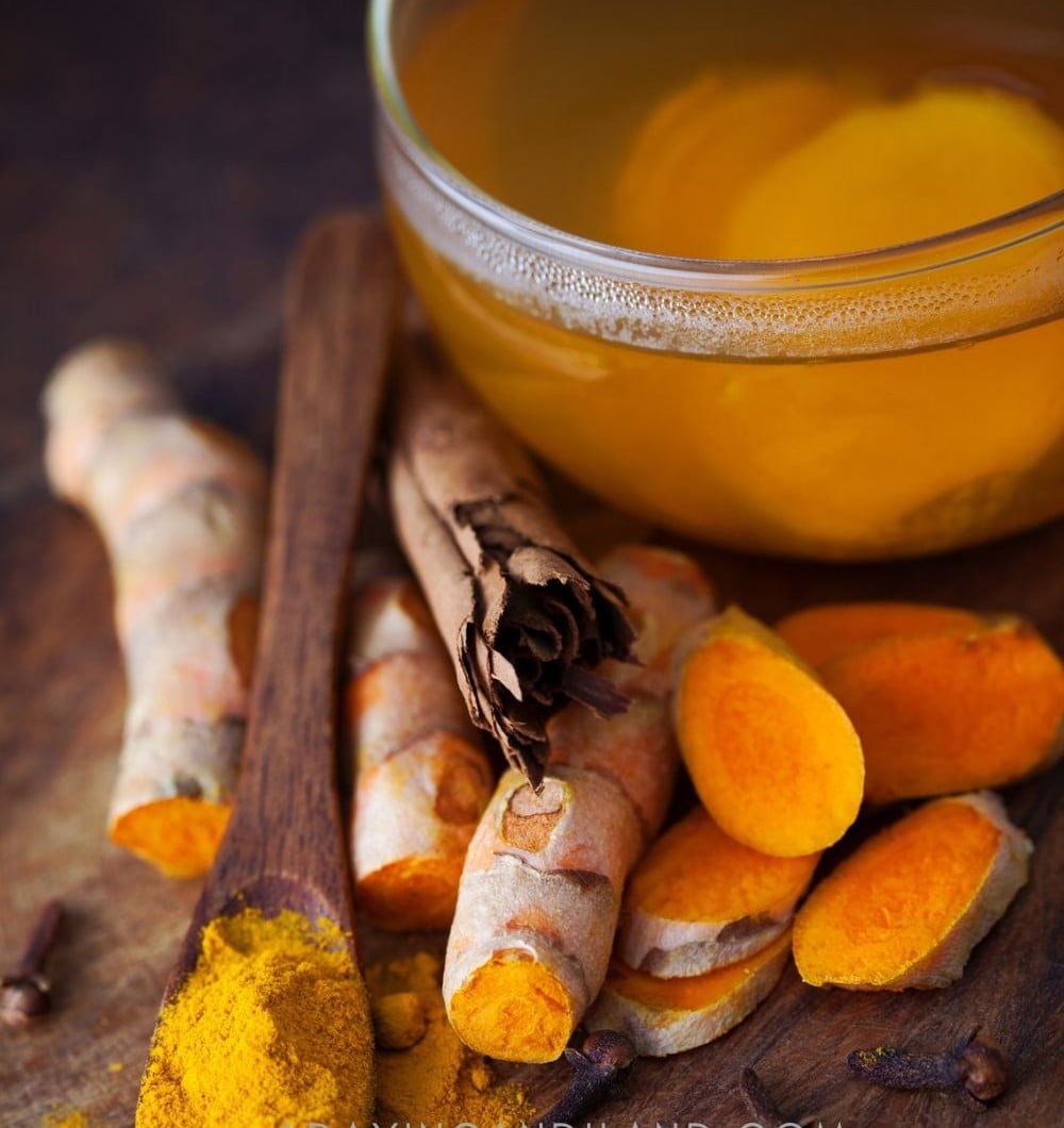 A glass mug of golden turmeric tea beside fresh turmeric root, cinnamon sticks and ground turmeric.