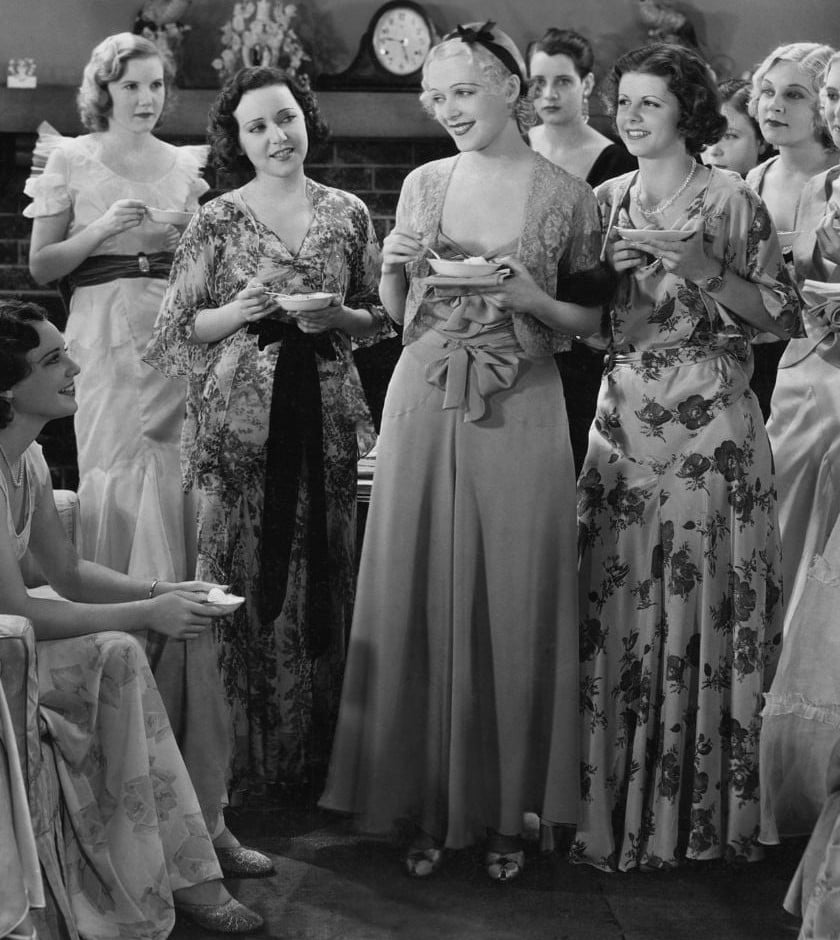 Women in long floral and satin dresses holding teacups and saucers at a vintage black and white tea party.